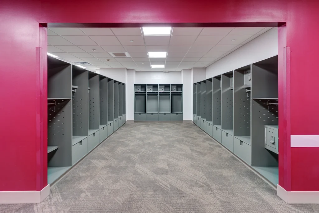 Empty gray locker room with open lockers along both walls and a bright pink entrance frame; fluorescent ceiling lights illuminate the space, part of exciting Campus Transformations, while the carpet features a neutral patterned design.