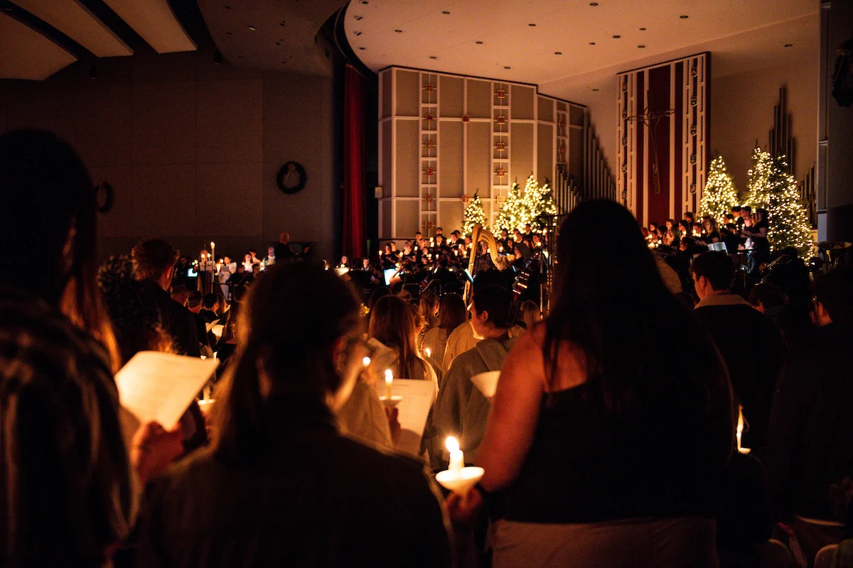 A large group of people hold lit candles and sheet music in a dimly lit church decorated with Christmas trees and lights, attending a holiday service or concert.