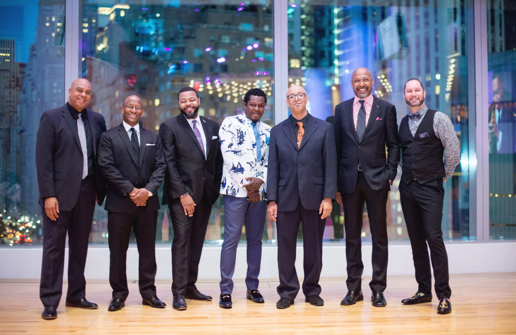 Seven men in suits and dress attire stand in a row, smiling, in front of large windows with a cityscape and colorful lights in the background.
