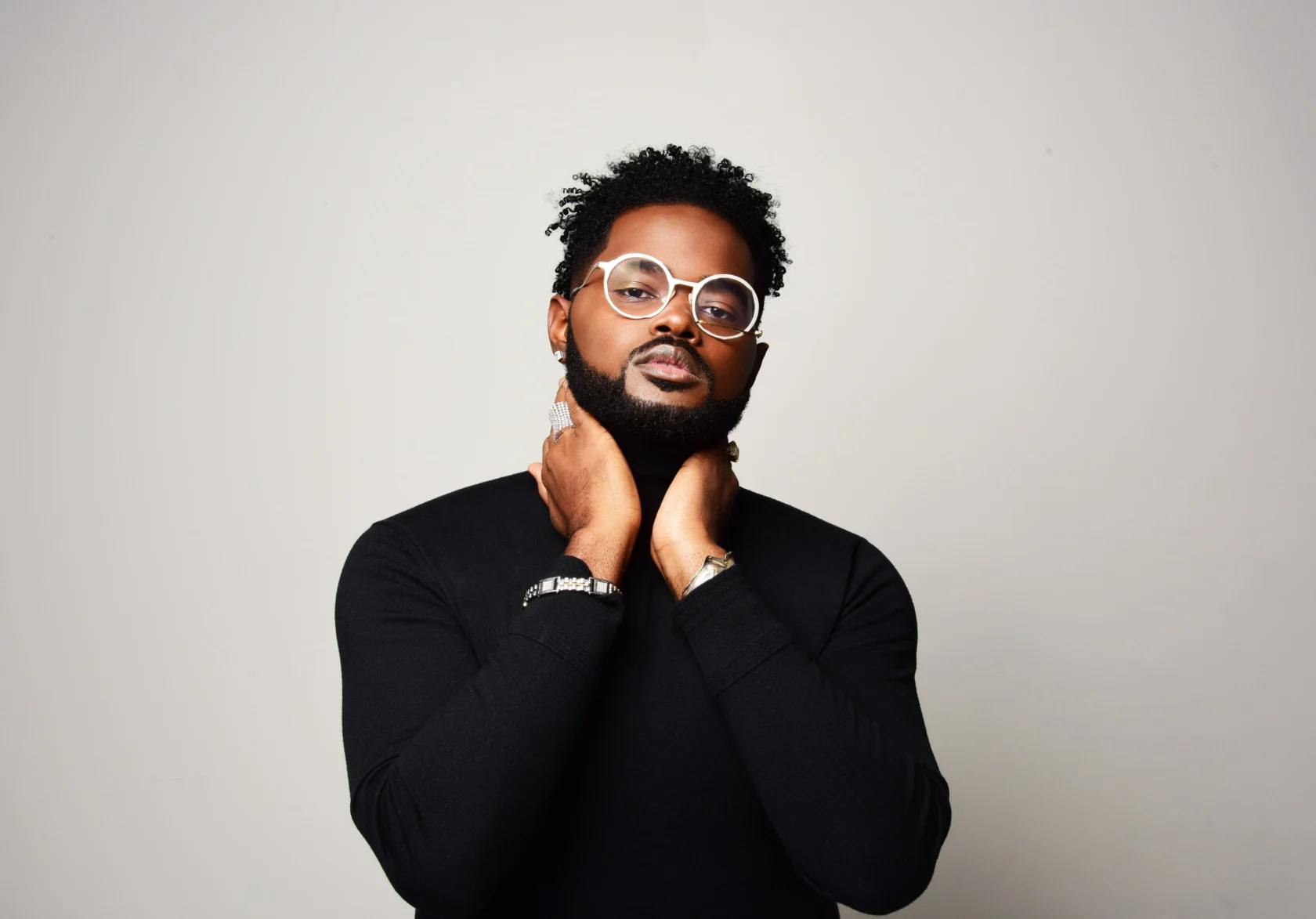 A man with a beard and short curly hair, wearing round glasses and a black long-sleeve shirt, stands against a plain light background with his hands gently crossed at his neck, looking confidently at the camera.
