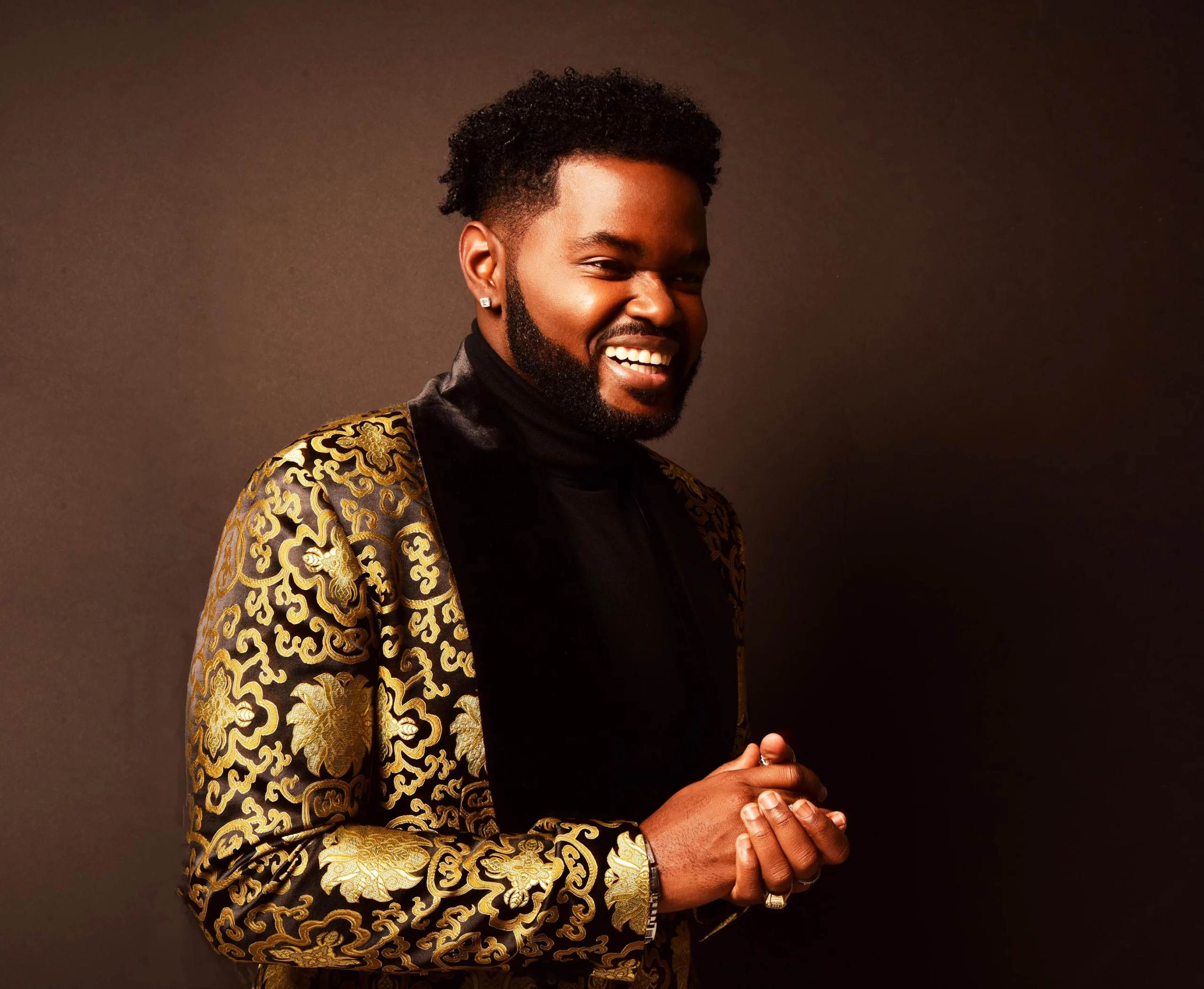 A man with a short curly hairstyle and beard smiles while wearing a black turtleneck and a gold-patterned blazer, standing against a dark background.
