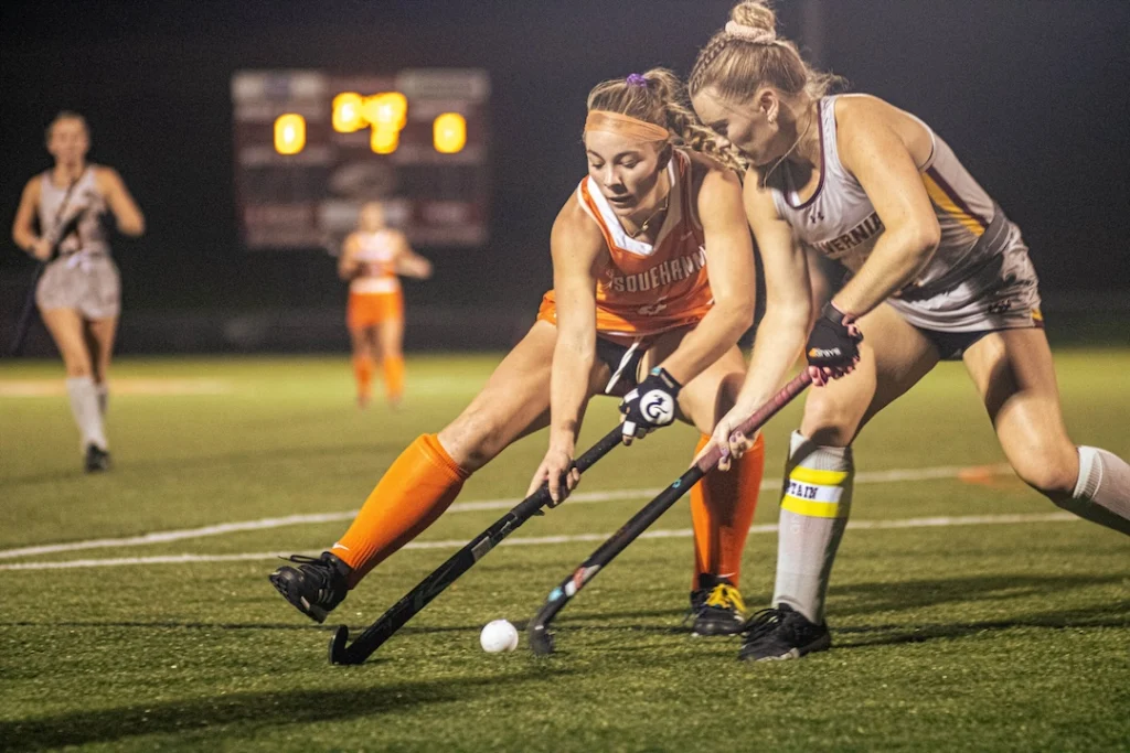 Two female field hockey players compete for the ball under stadium lights during a thrilling Homecoming-Reunion match. Both reach with their sticks, wearing orange and gray uniforms as the blurred scoreboard glows in the background.