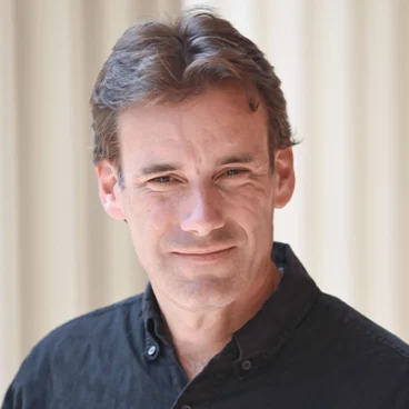 A man with short brown hair wearing a dark button-up shirt stands in front of a light, blurred background, looking at the camera and smiling slightly.