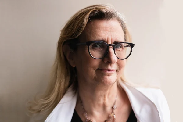 A middle-aged woman with long blonde hair and black-rimmed glasses smiles slightly while looking at the camera. She is wearing a white jacket over a black top and a beaded necklace. The background is neutral.