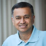 A middle-aged man with short dark hair and a slight smile is wearing a light blue polo shirt. He is posed in front of a neutral, softly blurred background.