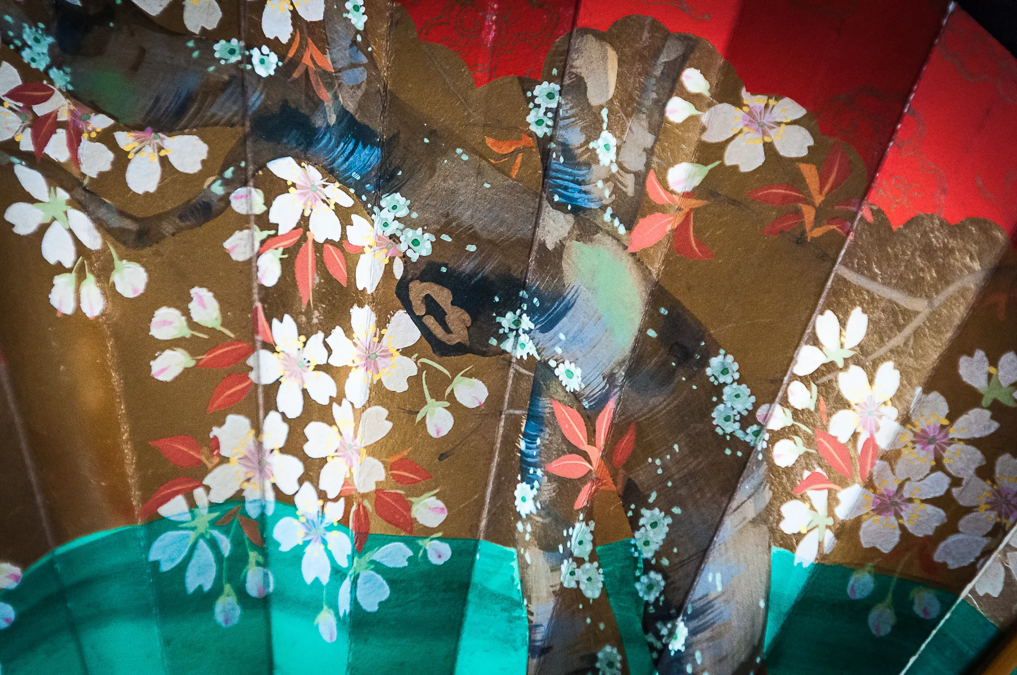 A close-up of a decorative fan painted with white cherry blossoms, a brown tree branch, and green and red accents on a gold background. The design features delicate floral details and vibrant colors.