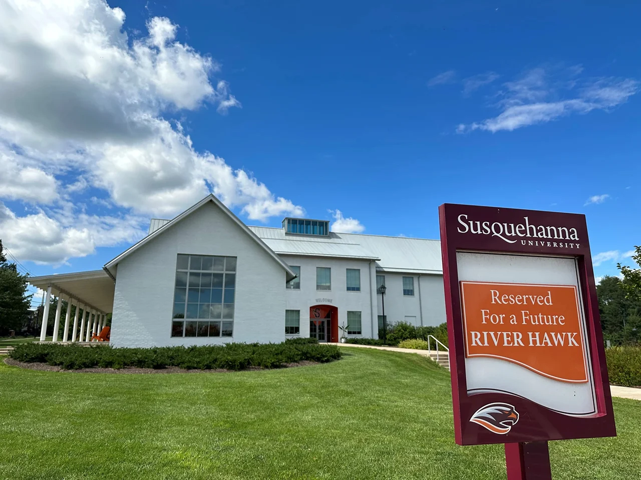 A white building with large windows stands under a blue sky with scattered clouds. In front, a red sign reads