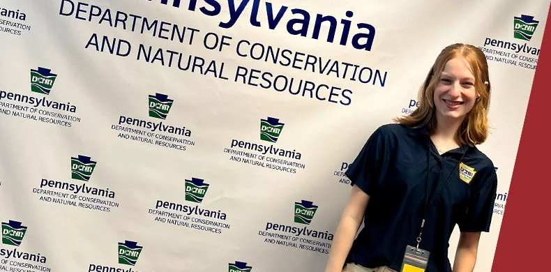 A young woman in a navy shirt and badge stands smiling in front of a Pennsylvania Department of Conservation and Natural Resources banner at an event.
