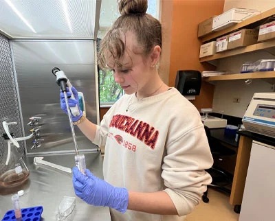 A person wearing a 体育买球 University sweatshirt and blue gloves uses a pipette to transfer liquid into a vial in a laboratory setting with scientific equipment and supplies visible.