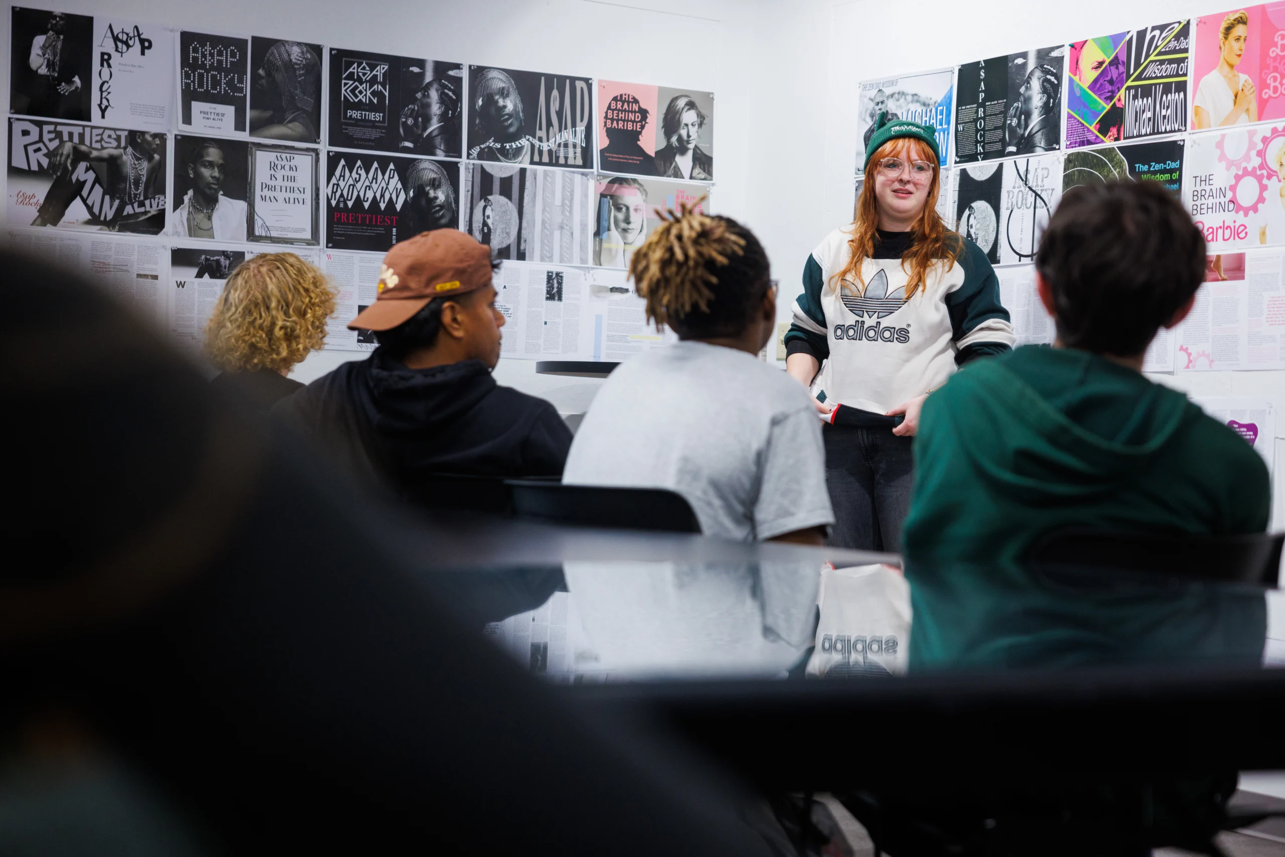 A woman in an Adidas sweatshirt and green beanie speaks to a group of seated students in a classroom with music posters and colorful images on the wall.