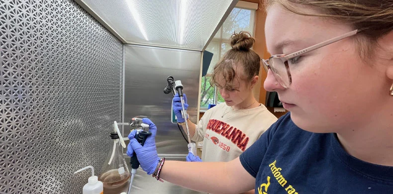 Two young women wearing gloves use pipettes to conduct an experiment in a laboratory fume hood. They are focused on their tasks, with laboratory equipment and bottles visible around them.