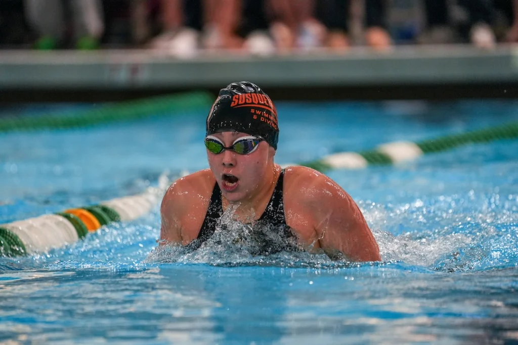 A swimmer wearing goggles and a black swim cap labeled “SUSQUEHANNA” competes in a breaststroke race during the Homecoming-Reunion, creating splashes with each stroke in the indoor pool.