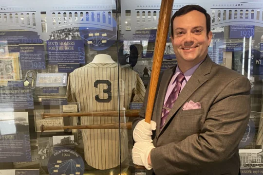 A man in a suit and white gloves smiles while holding a baseball bat in front of a display case with a vintage baseball jersey numbered 3 and various baseball memorabilia.