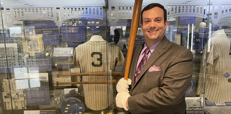 A man in a suit and white gloves smiles while holding a baseball bat in front of a display case with a vintage baseball jersey numbered 3 and various baseball memorabilia.