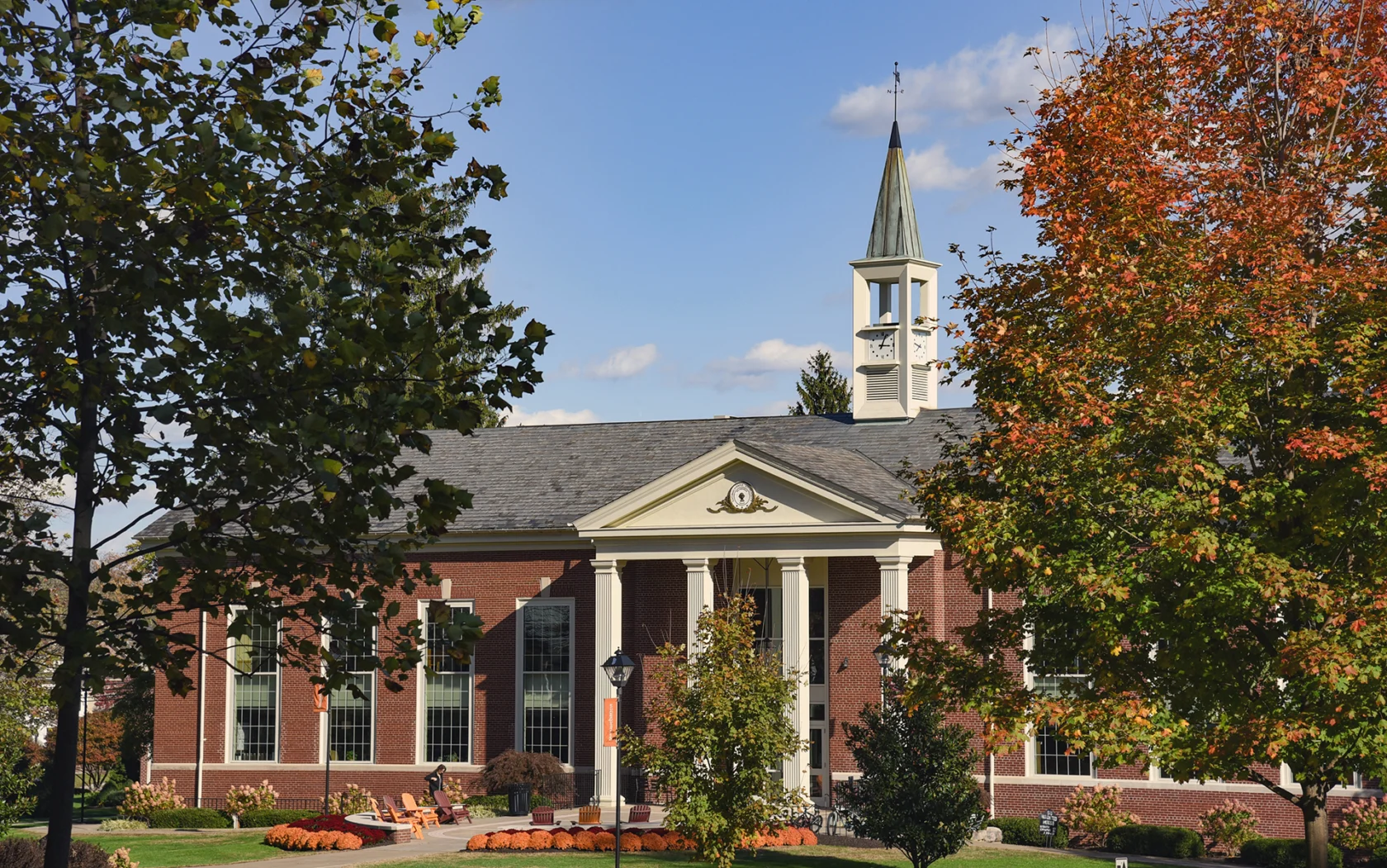 A red-brick building with white columns and a clock tower is surrounded by trees with green and orange leaves, under a blue sky with scattered clouds. This is the Blough-Weis Library.