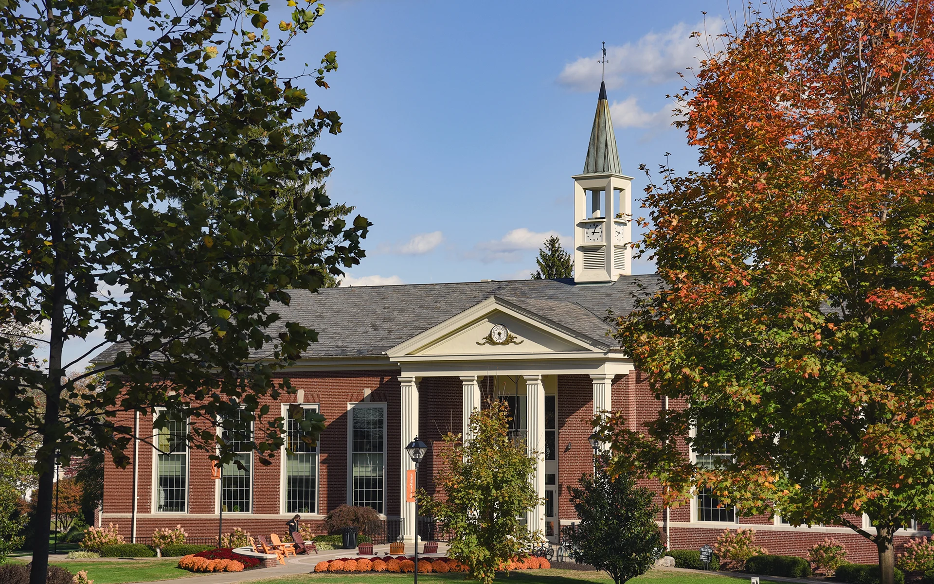 A red-brick building with white columns and a clock tower is surrounded by trees with green and orange leaves, under a blue sky with scattered clouds. This is the Blough-Weis Library.