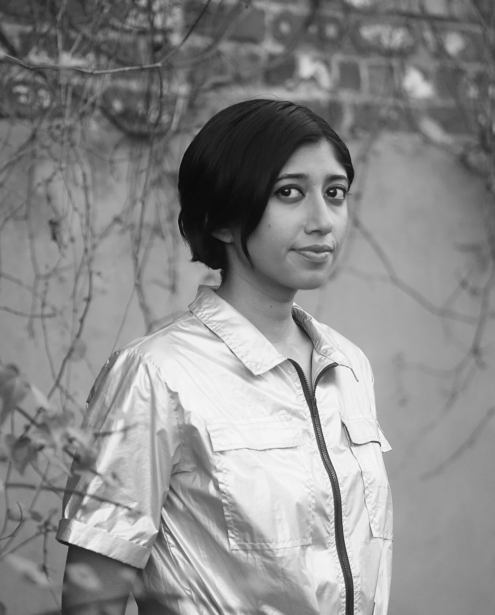 A person with short dark hair stands outdoors, wearing a shiny, light-colored zippered shirt. The background shows a wall with bare vines. This black and white photo was taken for the Seavey Reading Series.