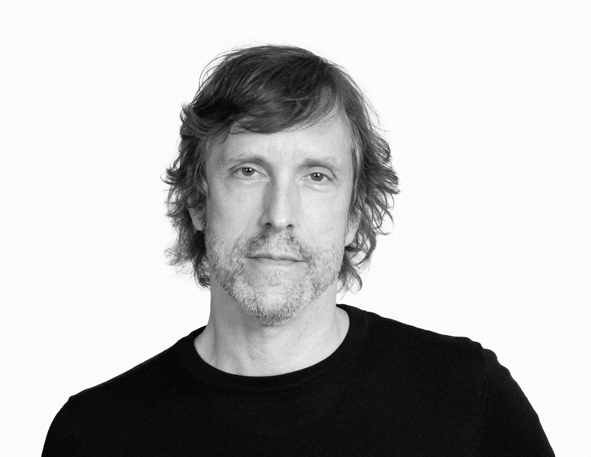 A middle-aged man with wavy, light brown hair and a short beard looks at the camera with a neutral expression. He is wearing a plain black shirt against a white background, ready to present at the Seavey Reading Series.