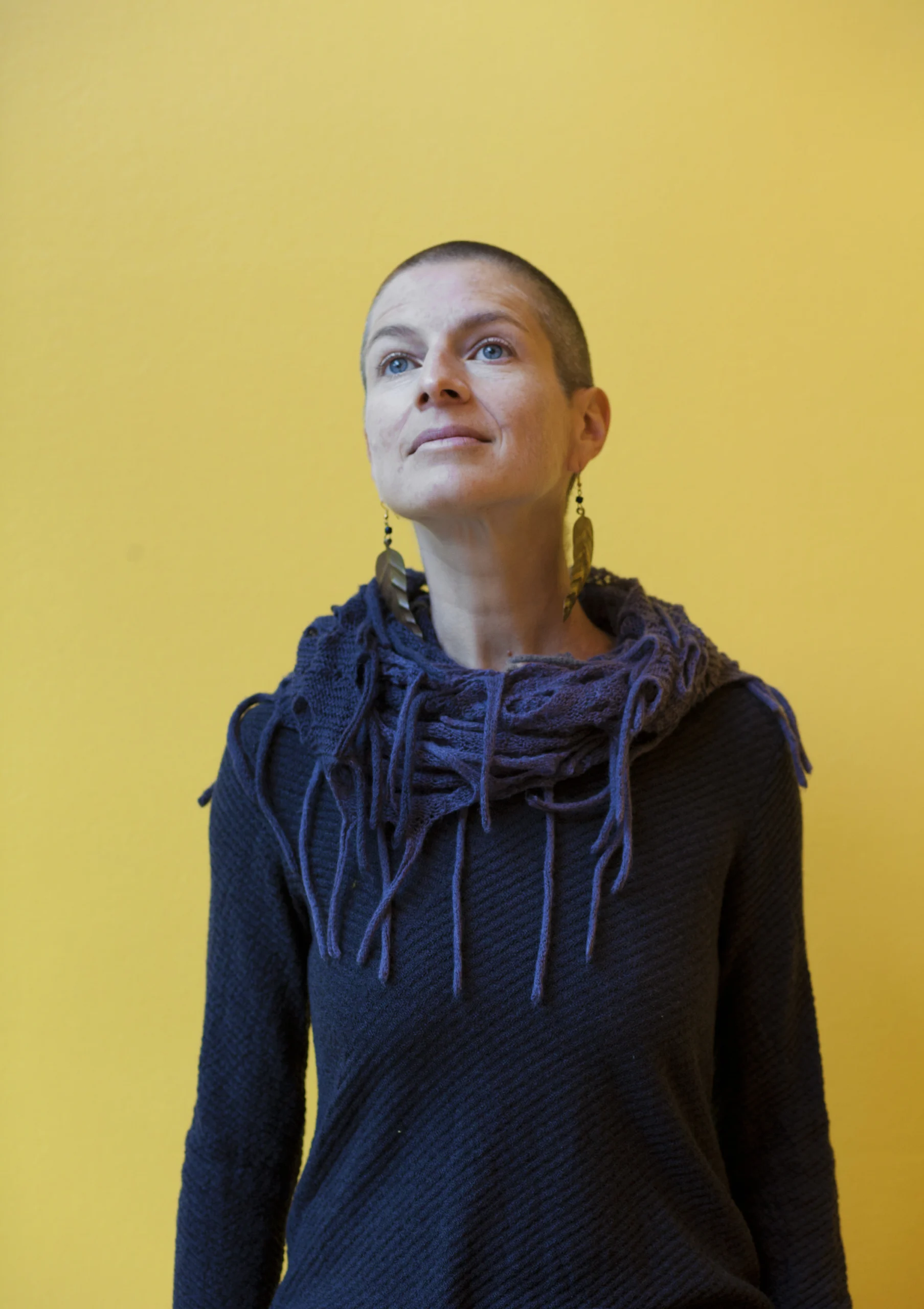 A person with a shaved head, wearing a textured dark sweater and a chunky scarf, stands in front of a solid yellow background, looking upward with a calm expression—a tranquil moment inspired by the Seavey Reading Series.
