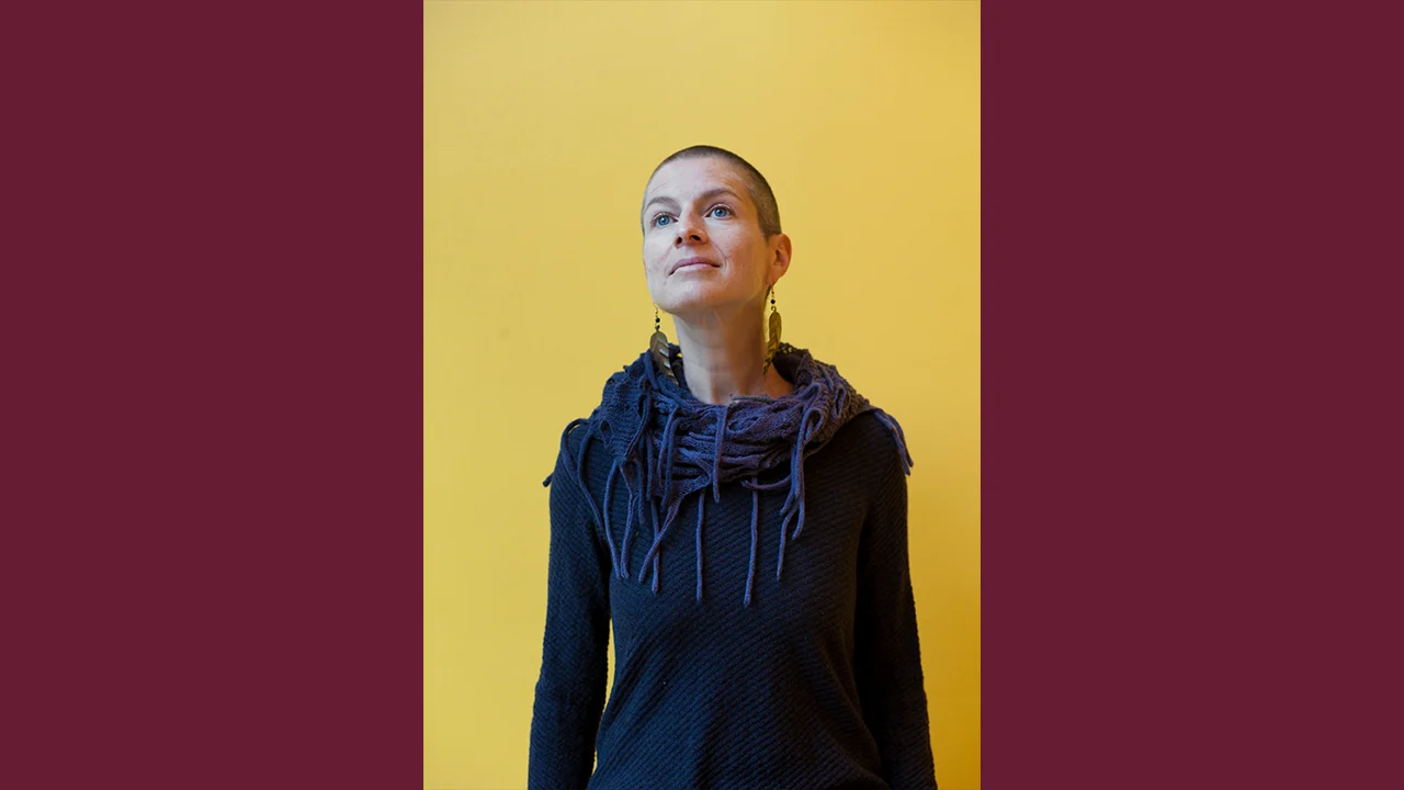 A person with a shaved head, wearing long earrings, a dark textured sweater, and a scarf, stands against a bright yellow background with maroon borders, looking slightly upward with a calm expression.