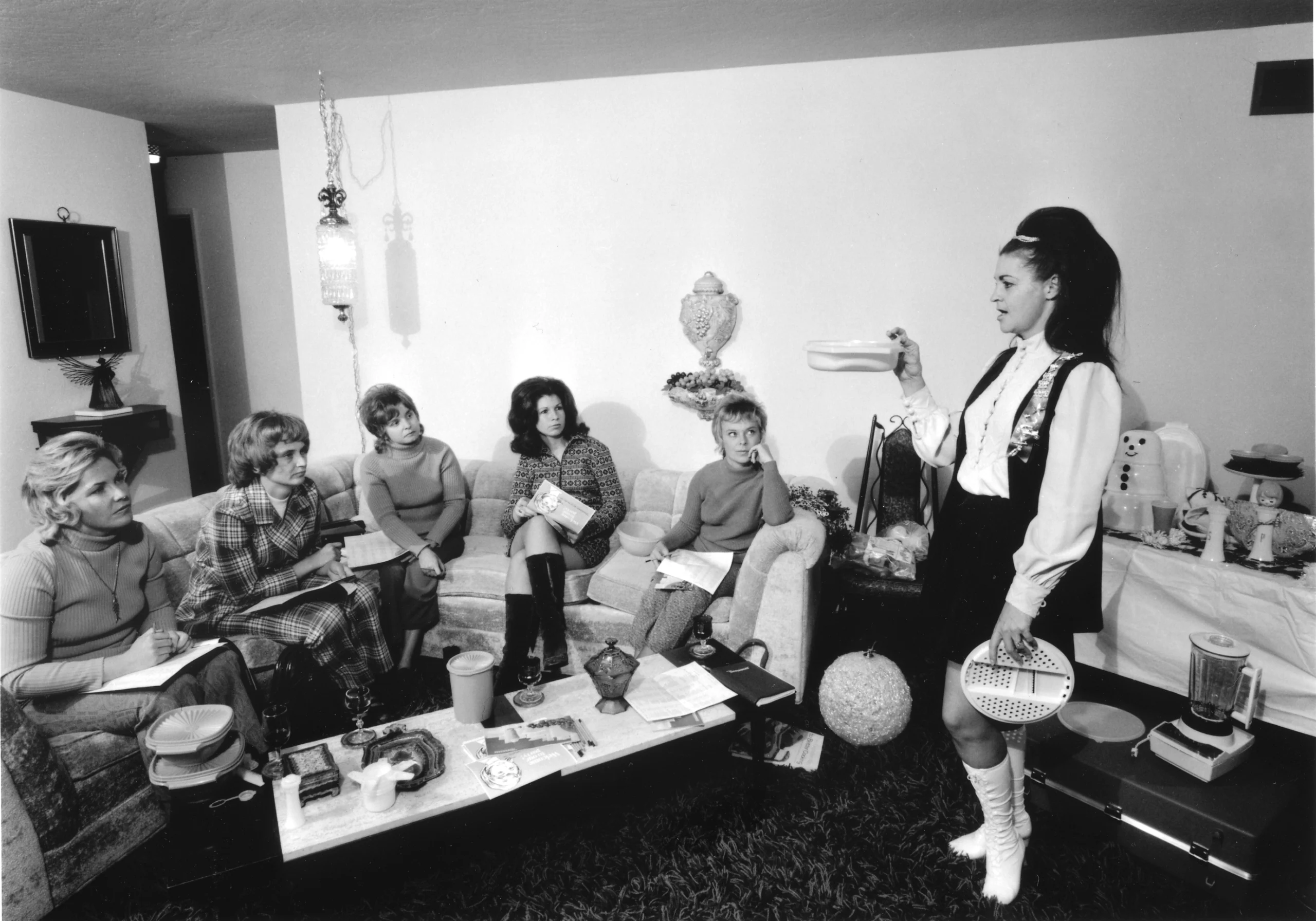 A woman stands holding a container, demonstrating a product to five seated women in a living room. The scene has a vintage feel, with 1970s outfits and home decor, and various items are displayed on tables.