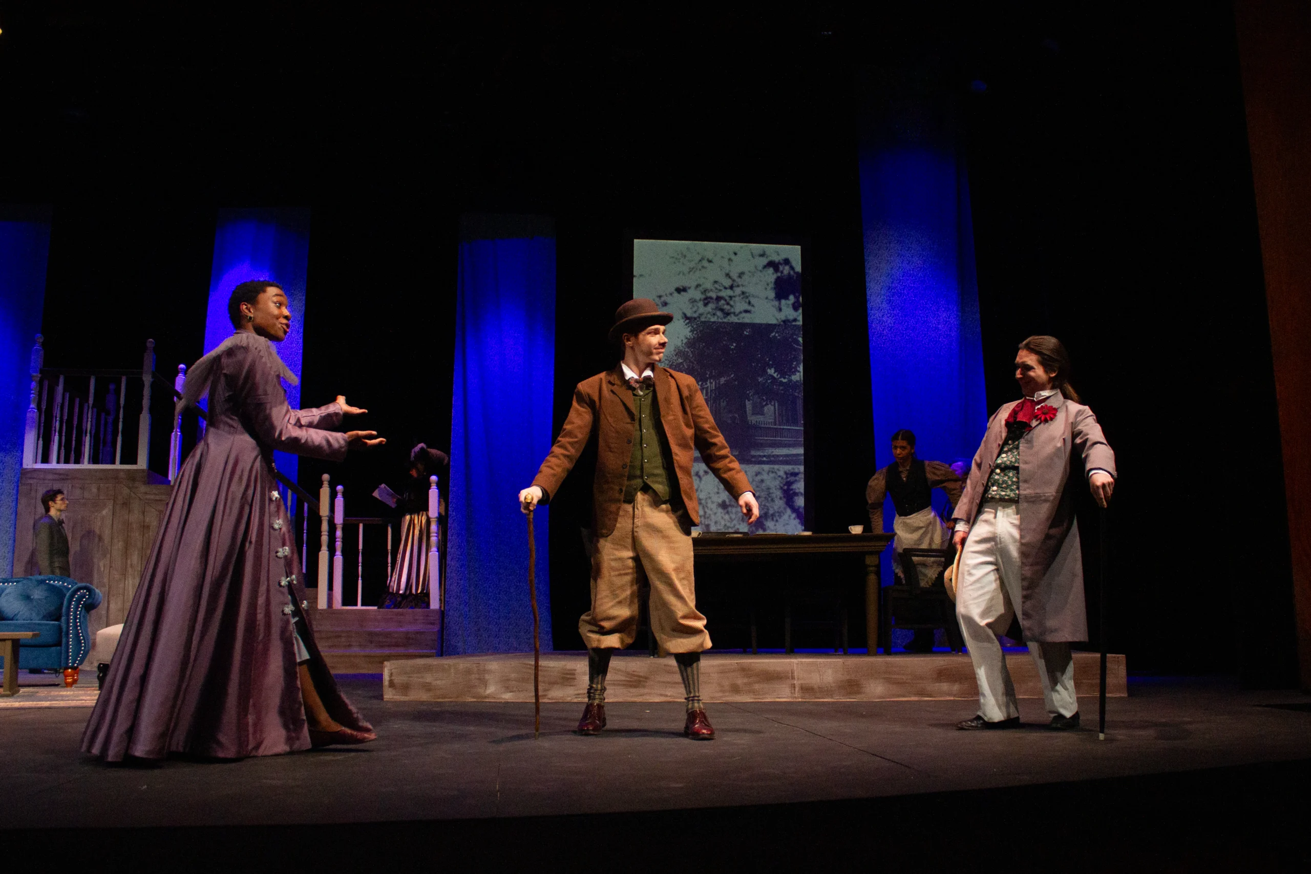 Three actors in period costumes perform on a stage set resembling an old house, with dramatic lighting and expressive gestures. Two other actors stand in the background, partially visible near a staircase and table.