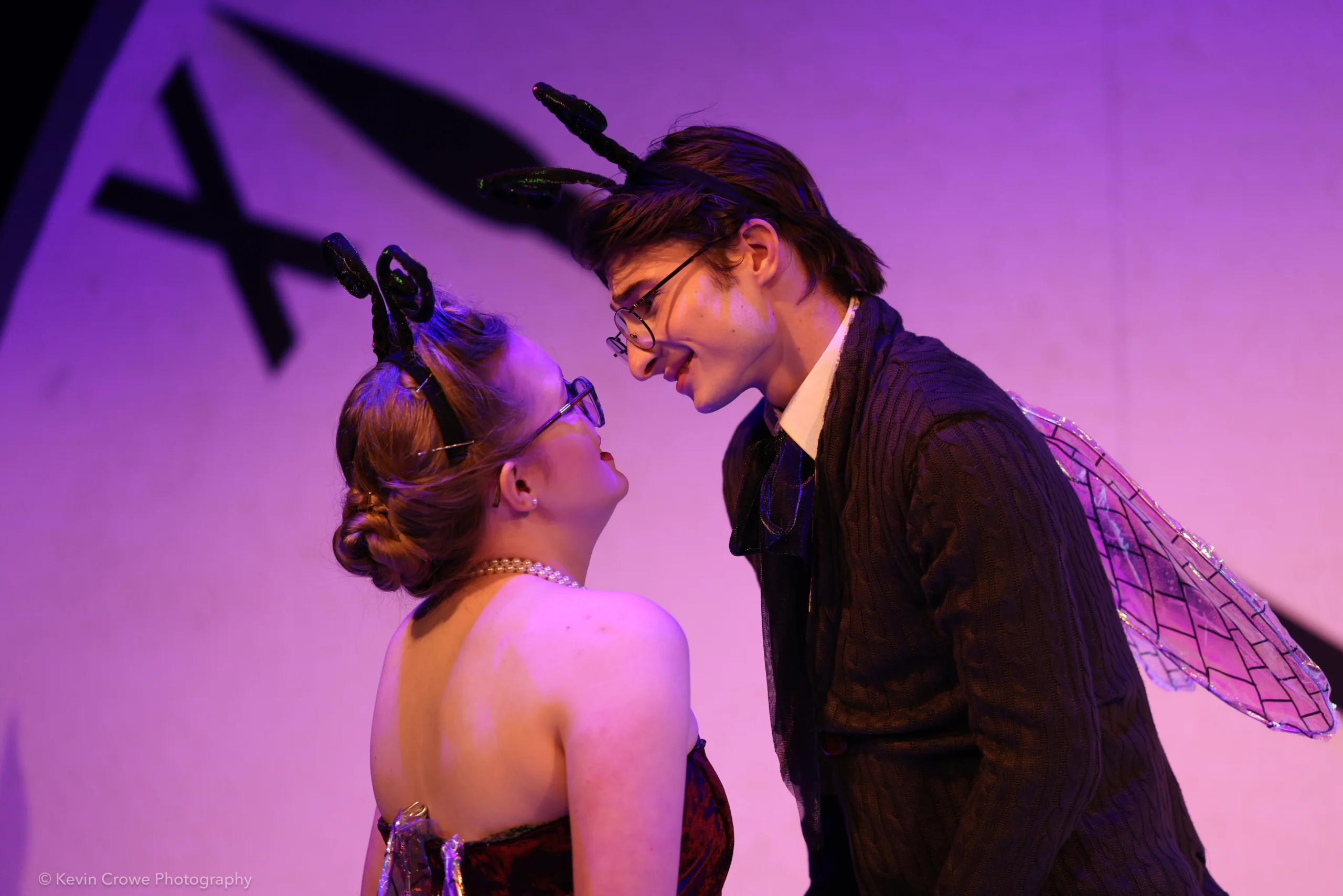 Two people dressed in costumes with black antennae headbands and clear wings face each other closely, smiling under purple lighting, suggesting a playful or affectionate interaction on stage.