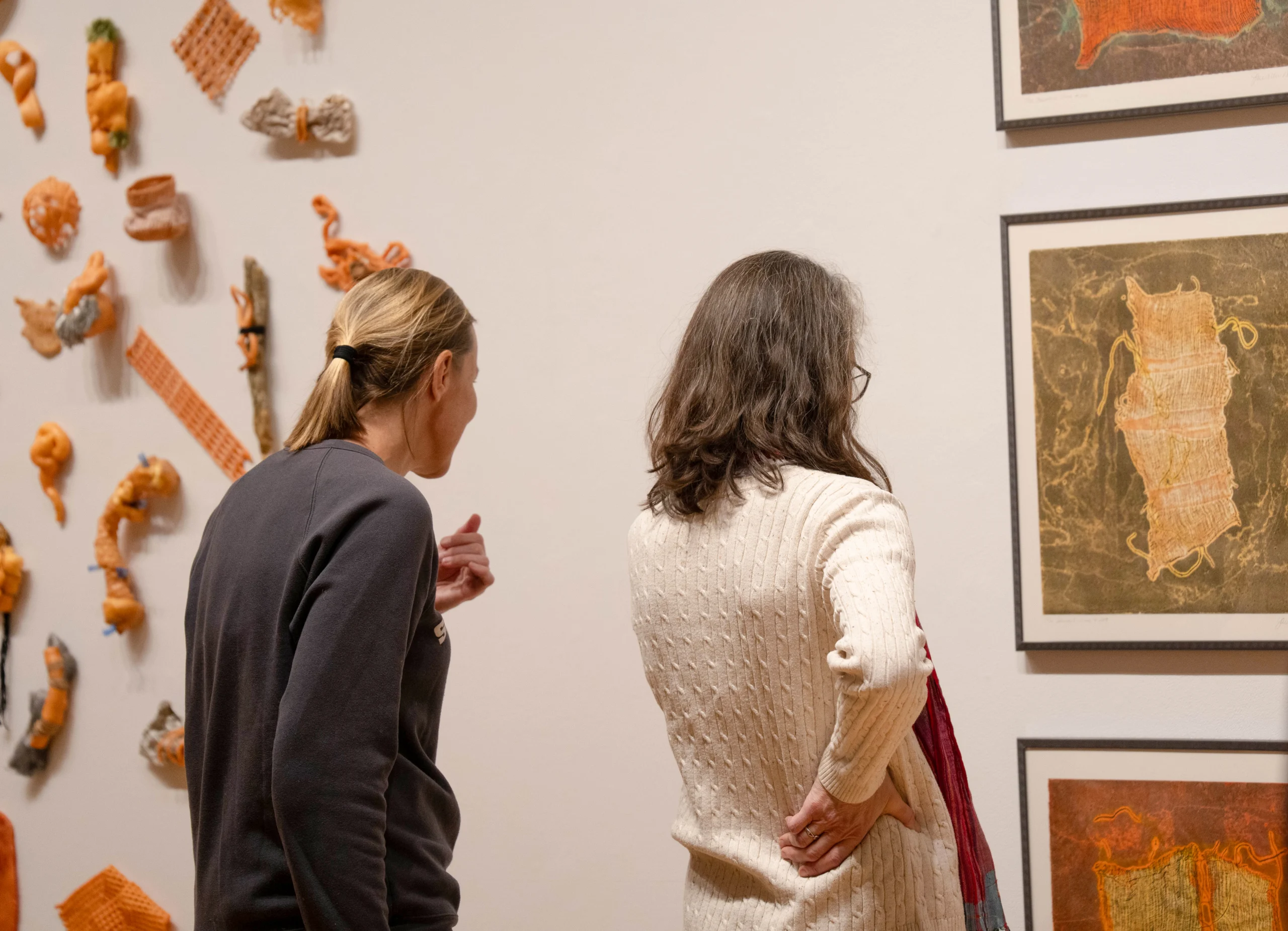 Two people stand in a gallery, observing framed abstract art on the wall. One person gestures while the other stands with a hand on their hip. Various orange sculptural pieces are also displayed on the adjacent wall.