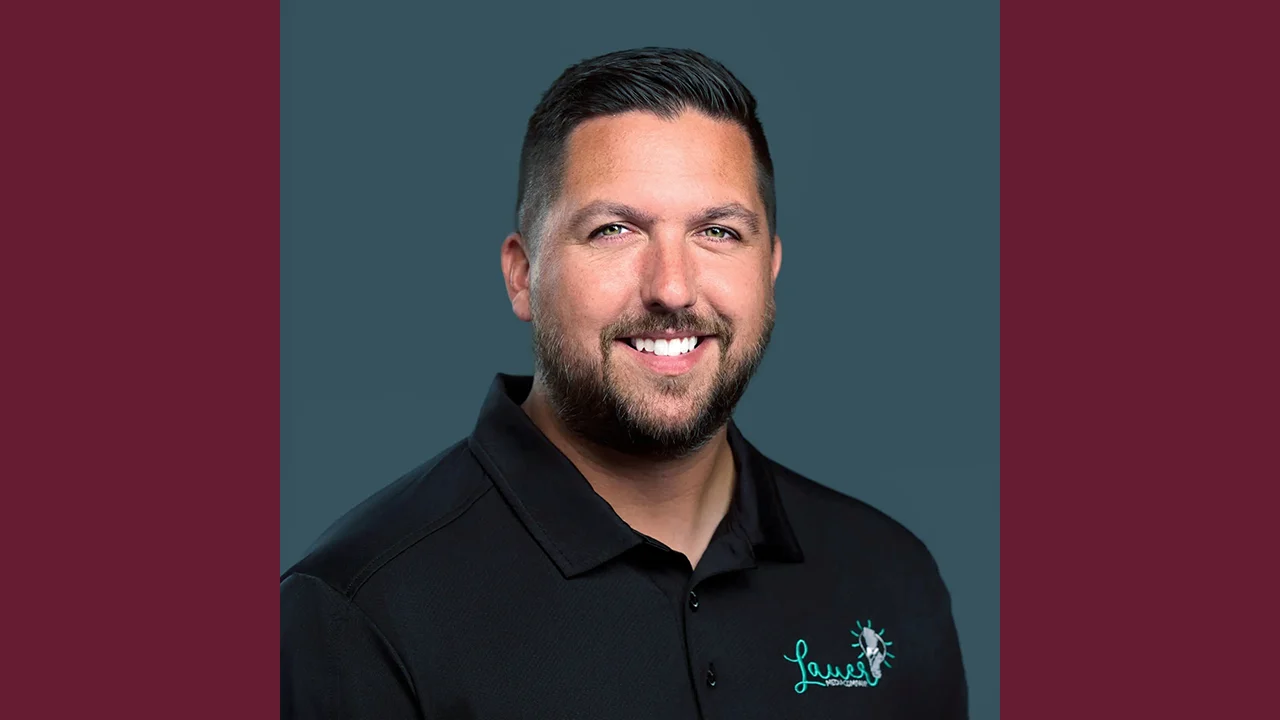 A man with short dark hair and a beard smiles at the camera. He is wearing a black collared shirt with a teal and white logo on the left side, and the background is solid dark blue with maroon borders on each side.