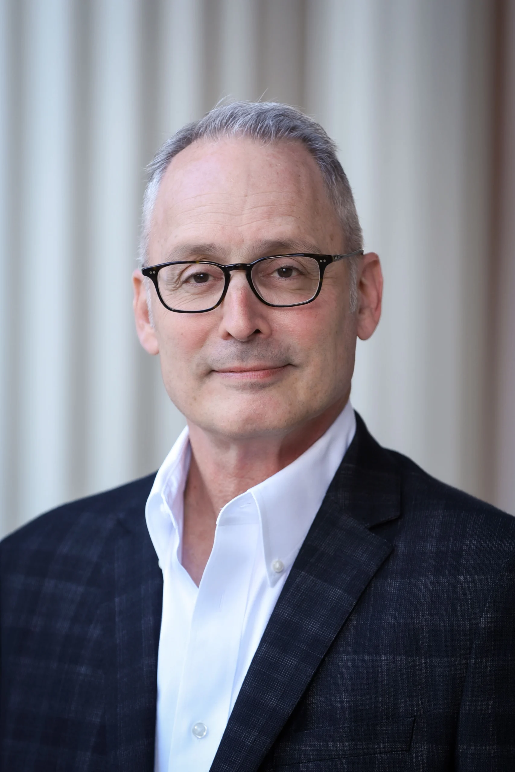 A middle-aged man with short gray hair and glasses, wearing a dark plaid blazer and white dress shirt, stands in front of a blurred, light-colored background, looking directly at the camera with a slight smile—perfect for the Seavey Reading Series.
