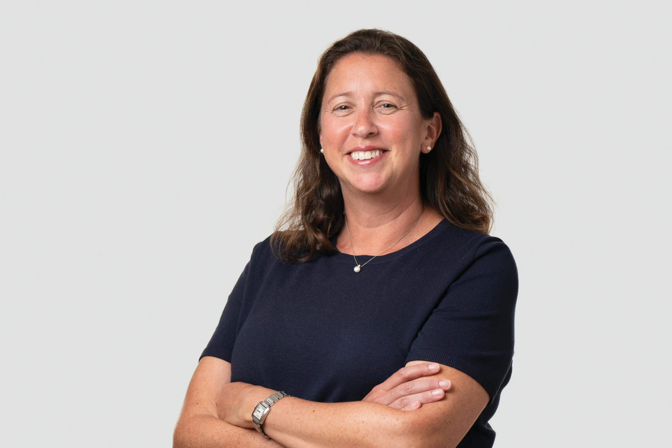 A woman with long brown hair wearing a dark short-sleeve top stands against a light background, smiling with her arms crossed. She is wearing a watch and a necklace.