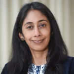 A woman with long black hair and dark eyes, wearing a dark blazer over a white top with blue patterns, poses in front of a light-colored, blurred background. She is smiling softly at the camera.