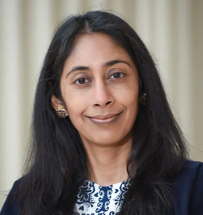 A woman with long black hair and dark eyes, wearing a dark blazer over a white top with blue patterns, poses in front of a light-colored, blurred background. She is smiling softly at the camera.