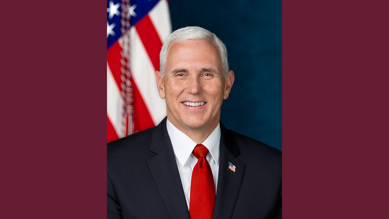 A man with short white hair in a dark suit, white shirt, and red tie smiles in front of an American flag and blue background.