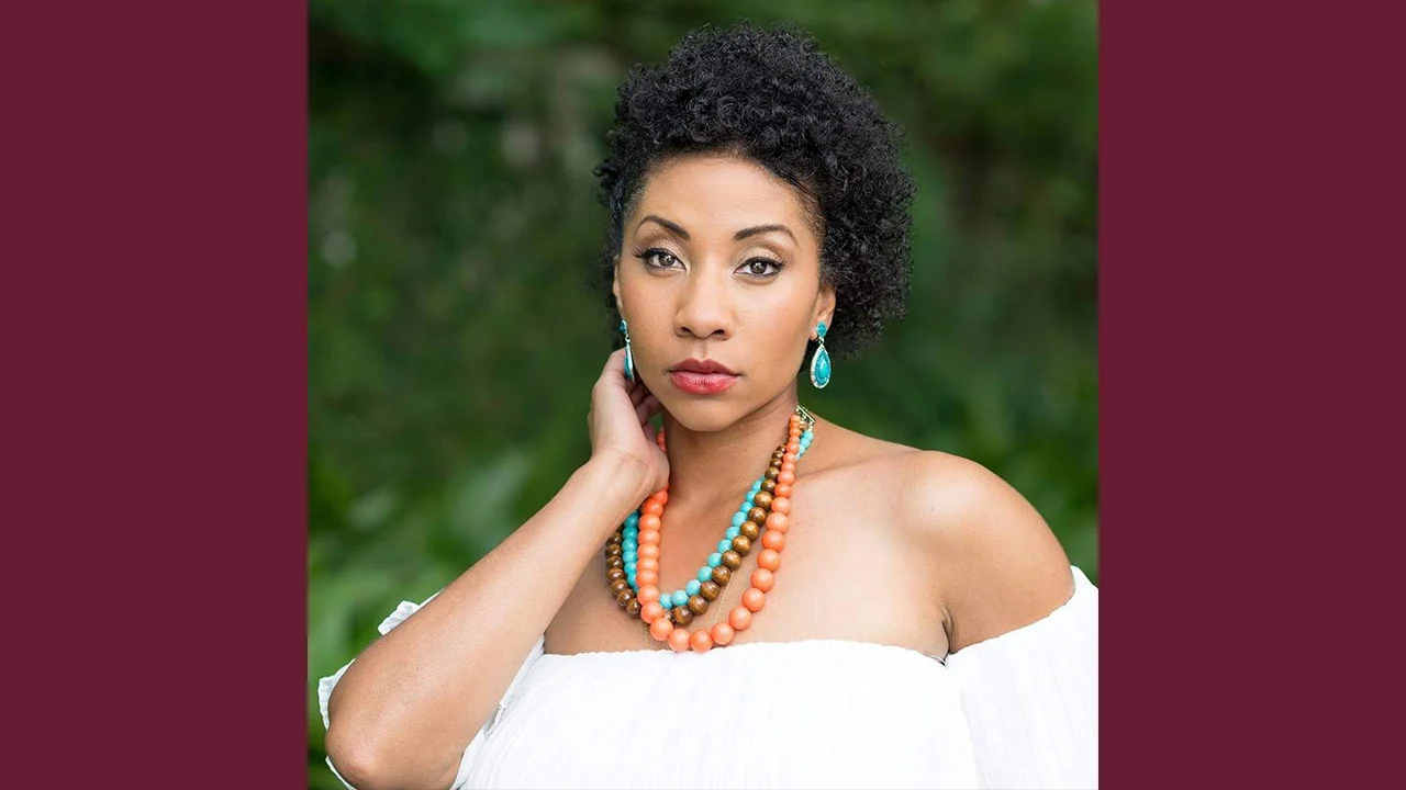 A woman with short curly hair wears a white off-the-shoulder top, turquoise earrings, and layered orange and turquoise necklaces, standing outdoors with greenery in the background. She looks at the camera, hand touching her neck.