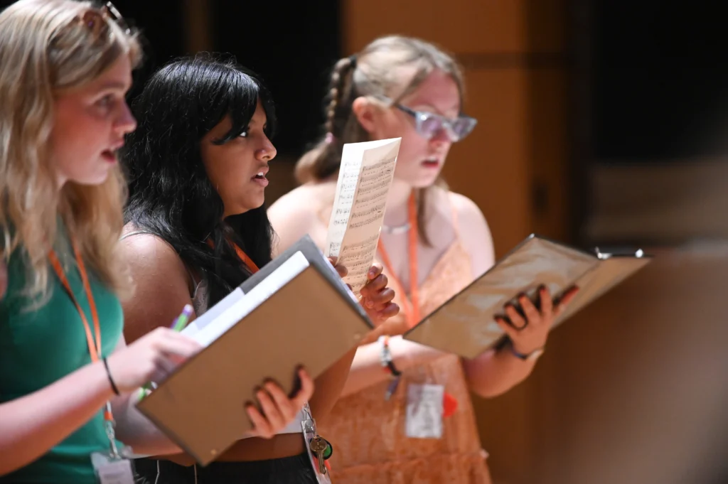 Three young women stand side by side, singing from music folders during a choir rehearsal—a scene reminiscent of Music Festivals & Workshops. Each wears a lanyard and casual summer clothing, fully focused on their sheet music.