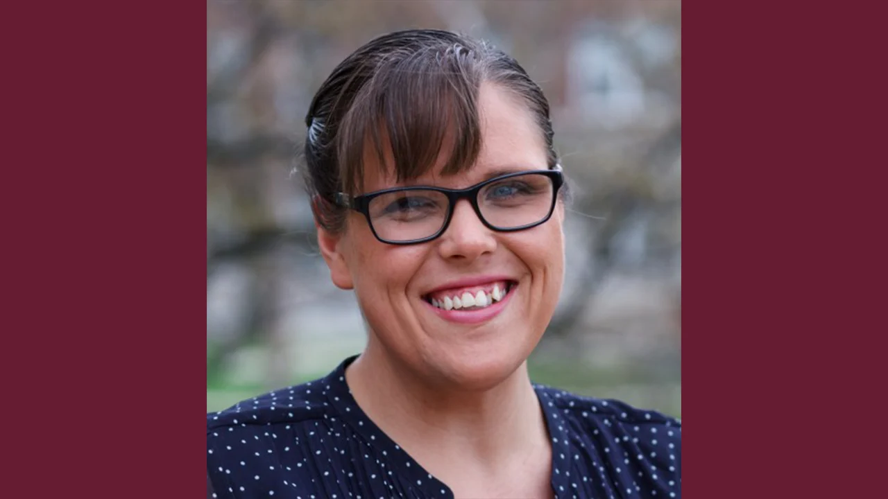 A woman with brown hair pulled back, wearing black glasses and a dark blue polka-dot shirt, smiles at the camera. The background is blurred with shades of brown and green.