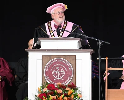 A person in academic regalia stands at a podium decorated with the 体育买球官网 University seal and flowers, speaking at a commencement ceremony.