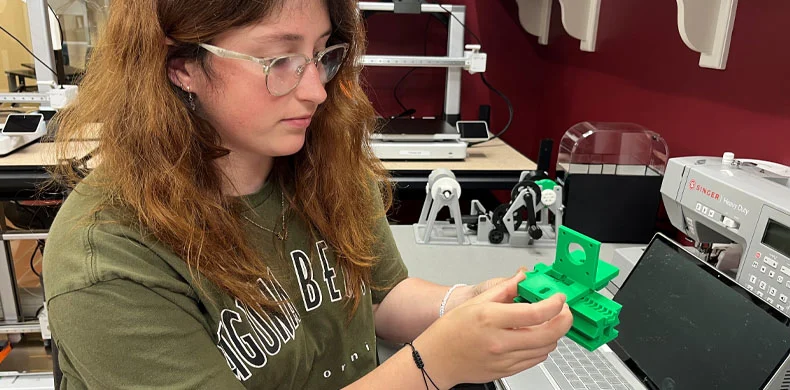 A person with long brown hair and glasses examines a green 3D-printed object in a laboratory setting, surrounded by equipment and a laptop.