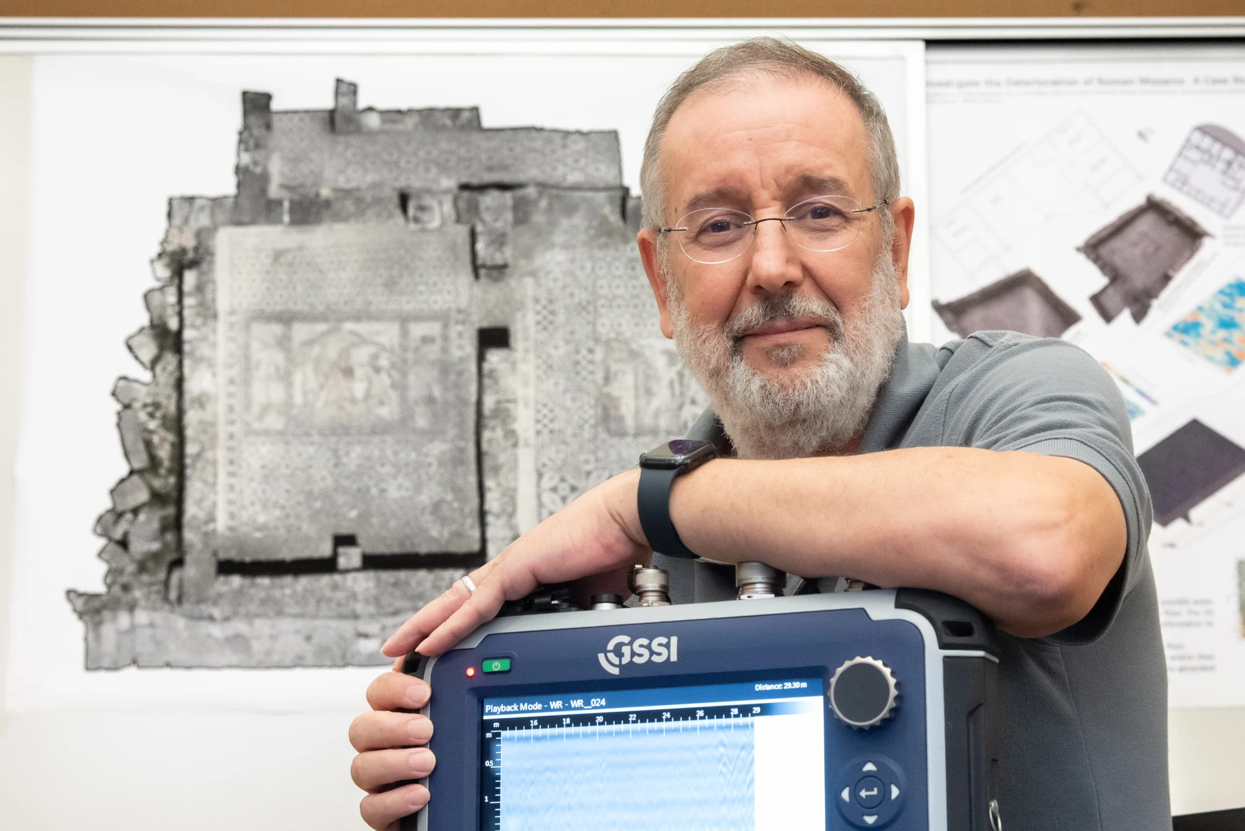 A man with glasses and a gray beard leans on a scientific instrument displaying a screen. Behind him, there is a large, detailed black-and-white archaeological image and diagrams pinned to the wall.