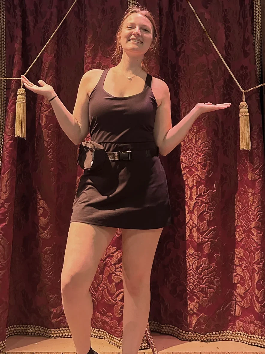 A woman in a black dress stands smiling with arms raised in front of ornate red curtains with gold tassels, set against a rustic wooden stage background.