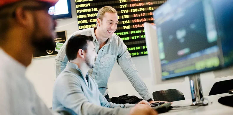 Three men in a modern office work at computer monitors, with stock market data displayed on screens in the background. One man stands and smiles while talking to two others who are seated and focused on their screens.