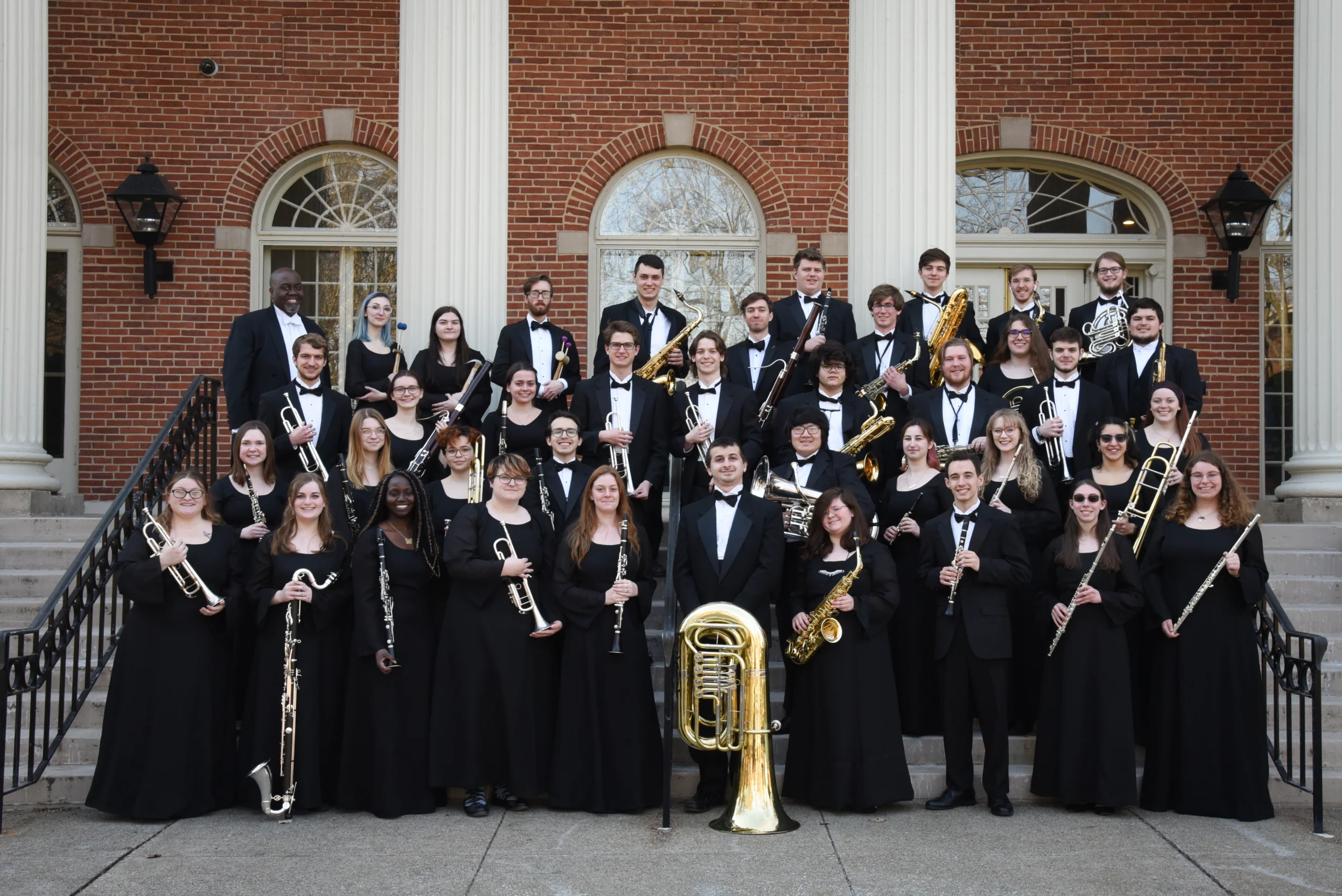 A large group of musicians in formal black attire stand together on steps in front of a brick building with columns, holding wind and brass instruments, smiling for a group photo at the Symposium on Historic American Music.