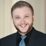 A young man with short light brown hair, a beard, and light eyes smiles at the camera. He is wearing a black suit jacket, black shirt, and a light blue tie. The background is softly blurred.