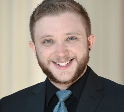 A young man with short light brown hair, a beard, and light eyes smiles at the camera. He is wearing a black suit jacket, black shirt, and a light blue tie. The background is softly blurred.