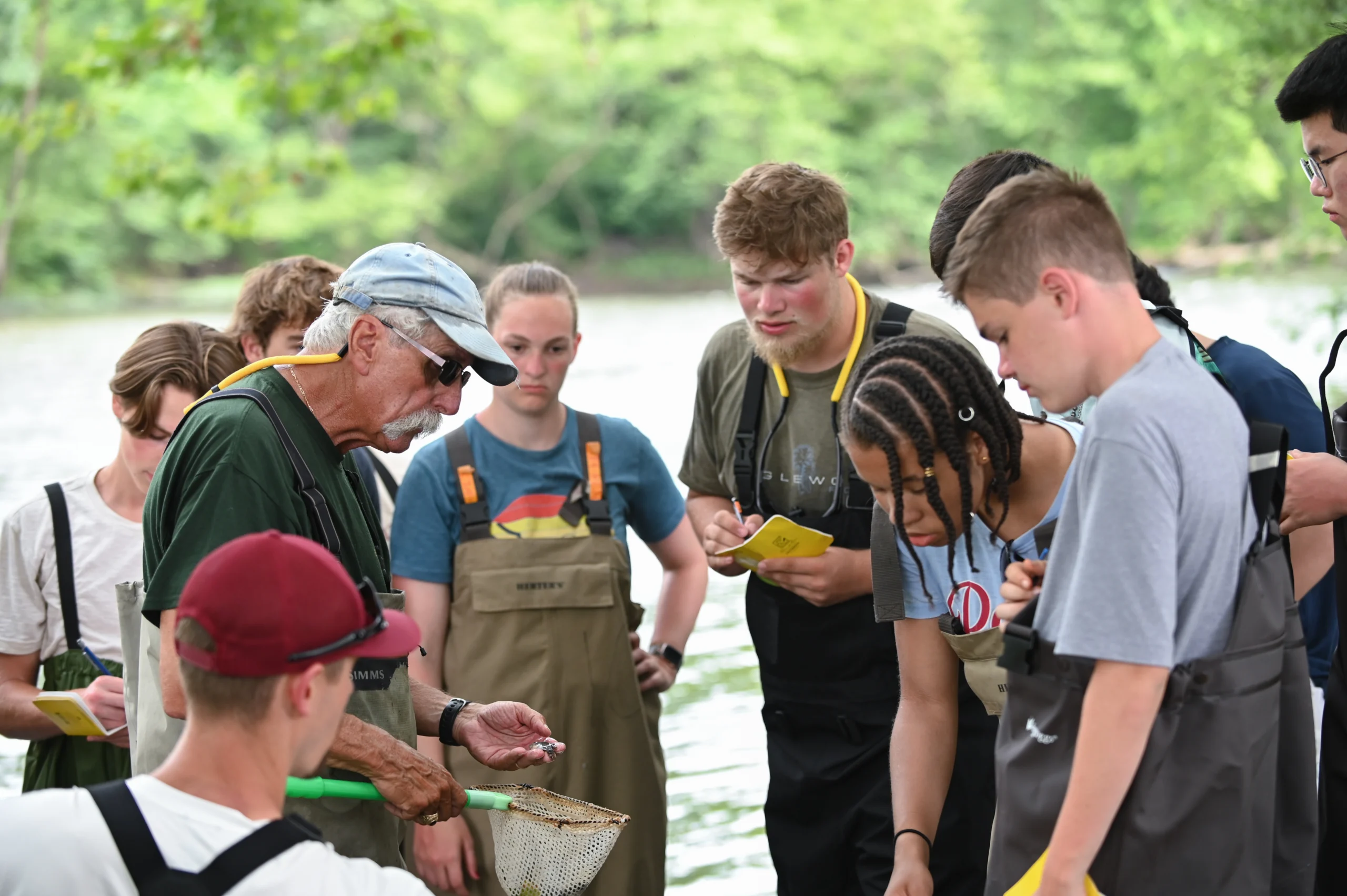 A group of young people in a Summer Pre-College Program and an older man in waders examine something small by a river. Focused, some hold notebooks as he shows a small object over a net, with trees and water visible in the background.