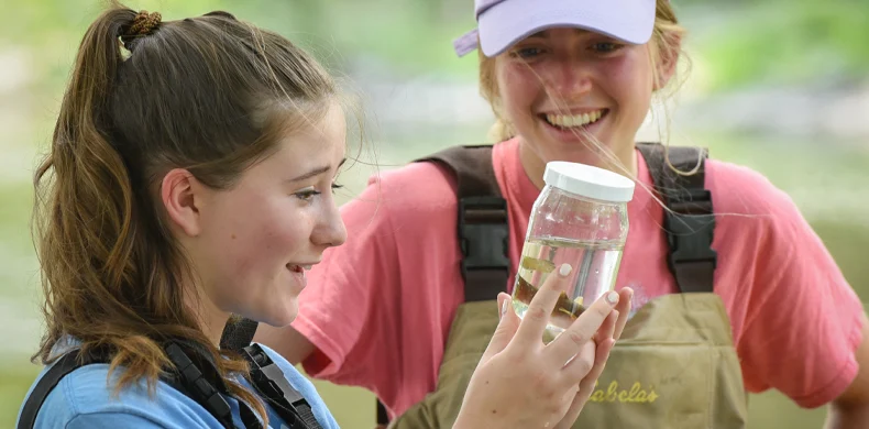 Two young women outdoors, wearing waders and smiling. One holds a clear jar filled with water and examines its contents closely, while the other looks on happily. Green, blurred background suggests a natural setting.
