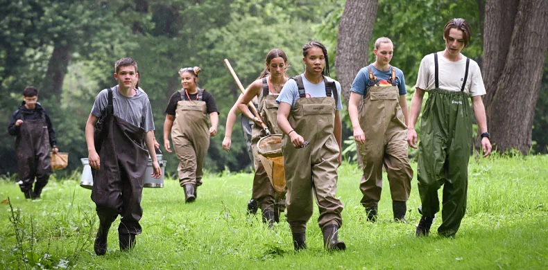 A group of young people in waders walk through a grassy area in a forest, carrying buckets and nets, possibly heading to a stream or river for a scientific or environmental activity.