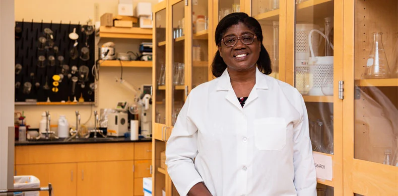 A woman wearing a white lab coat stands smiling in a laboratory filled with glassware, scientific equipment, and wooden cabinets.