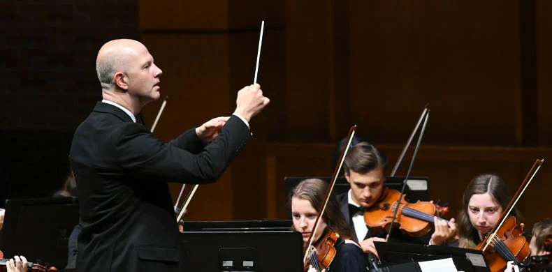A conductor in a black suit leads an orchestra, raising a baton, while young musicians play violins and other string instruments on stage.